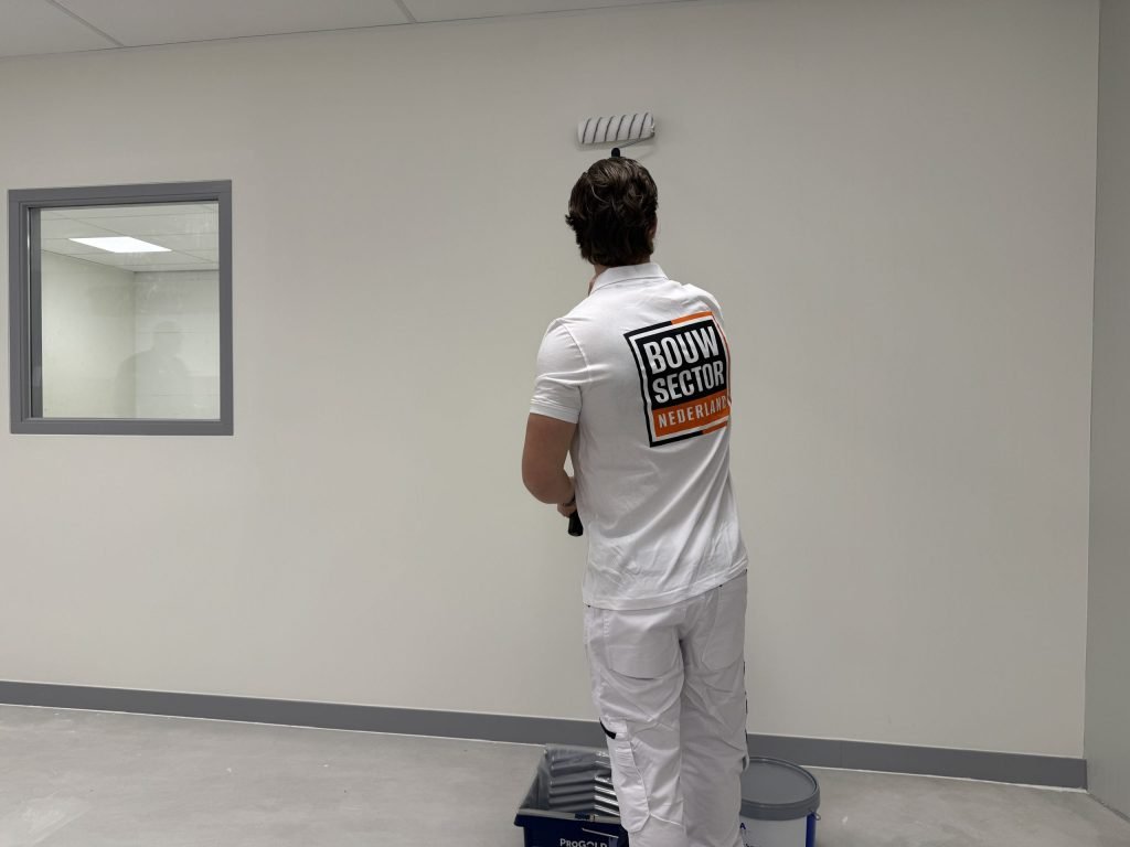 Schilder van Bouwsector Nederland rolt een lichte binnenmuur met muurverf, verfbak en emmer aan zijn voeten