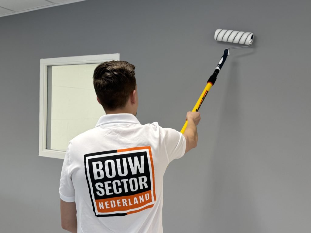 Schilder in wit werkshirt saust een grijze muur met een roller aan een verlengsteel naast een raam