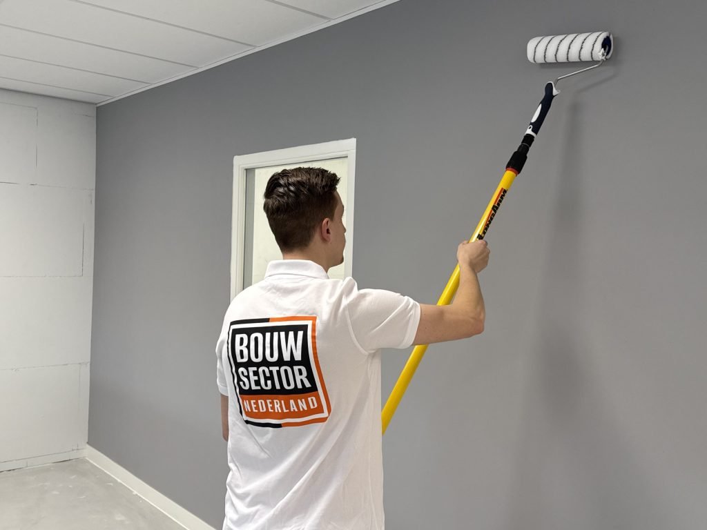 Grijze muur wordt tot tegen het plafond gesausd door een schilder met een lange verfrolle