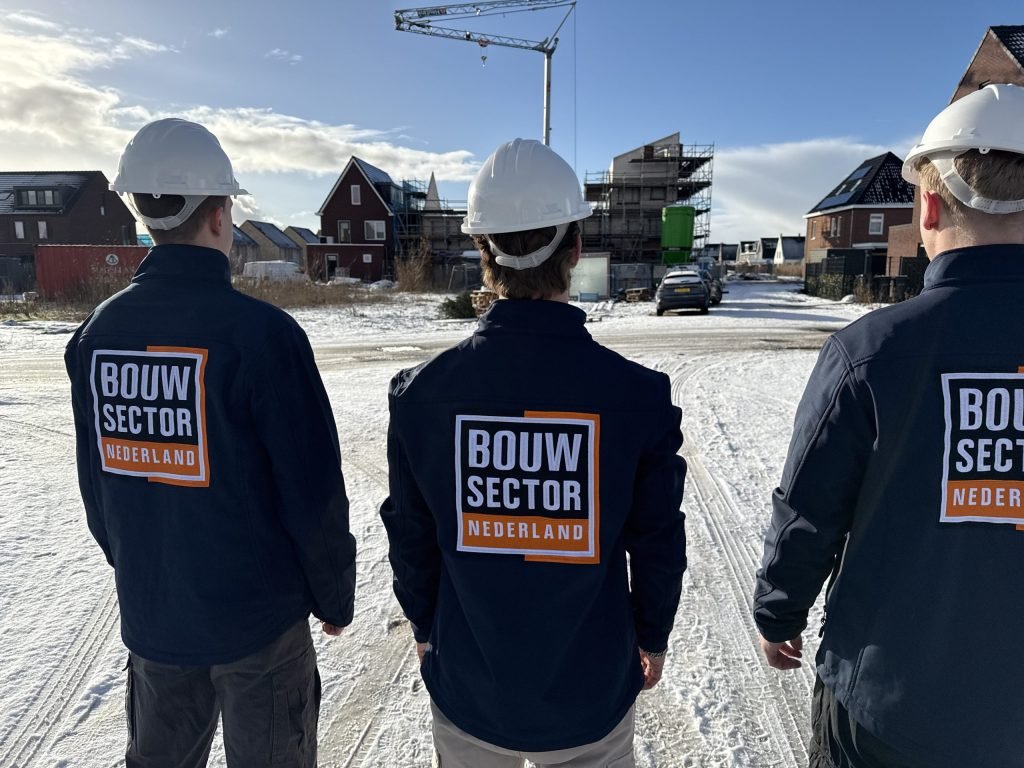 Drie vakmensen van Bouwsector Nederland met witte helm lopen over een besneeuwde straat naar woningen in aanbouw