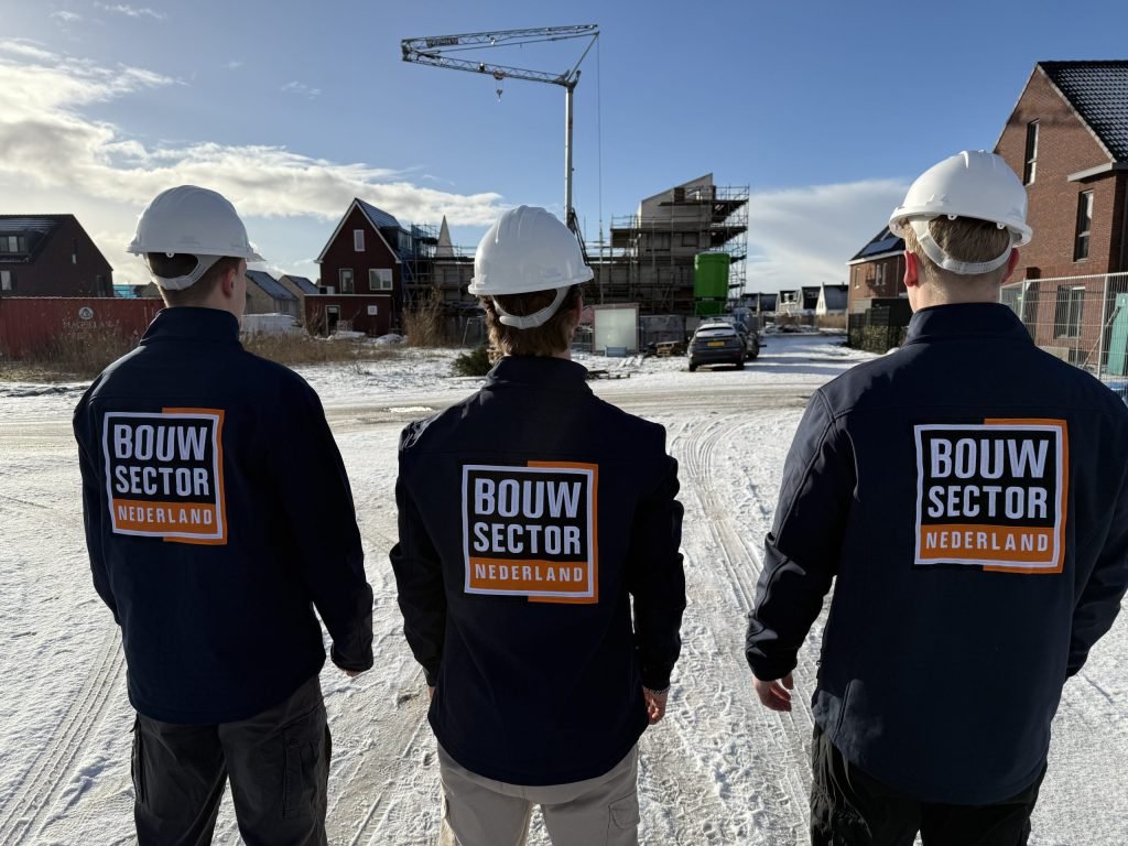 Drie vakmensen van Bouwsector Nederland met witte helm kijken vanaf een besneeuwde weg naar nieuwe huizen en een bouwkraan